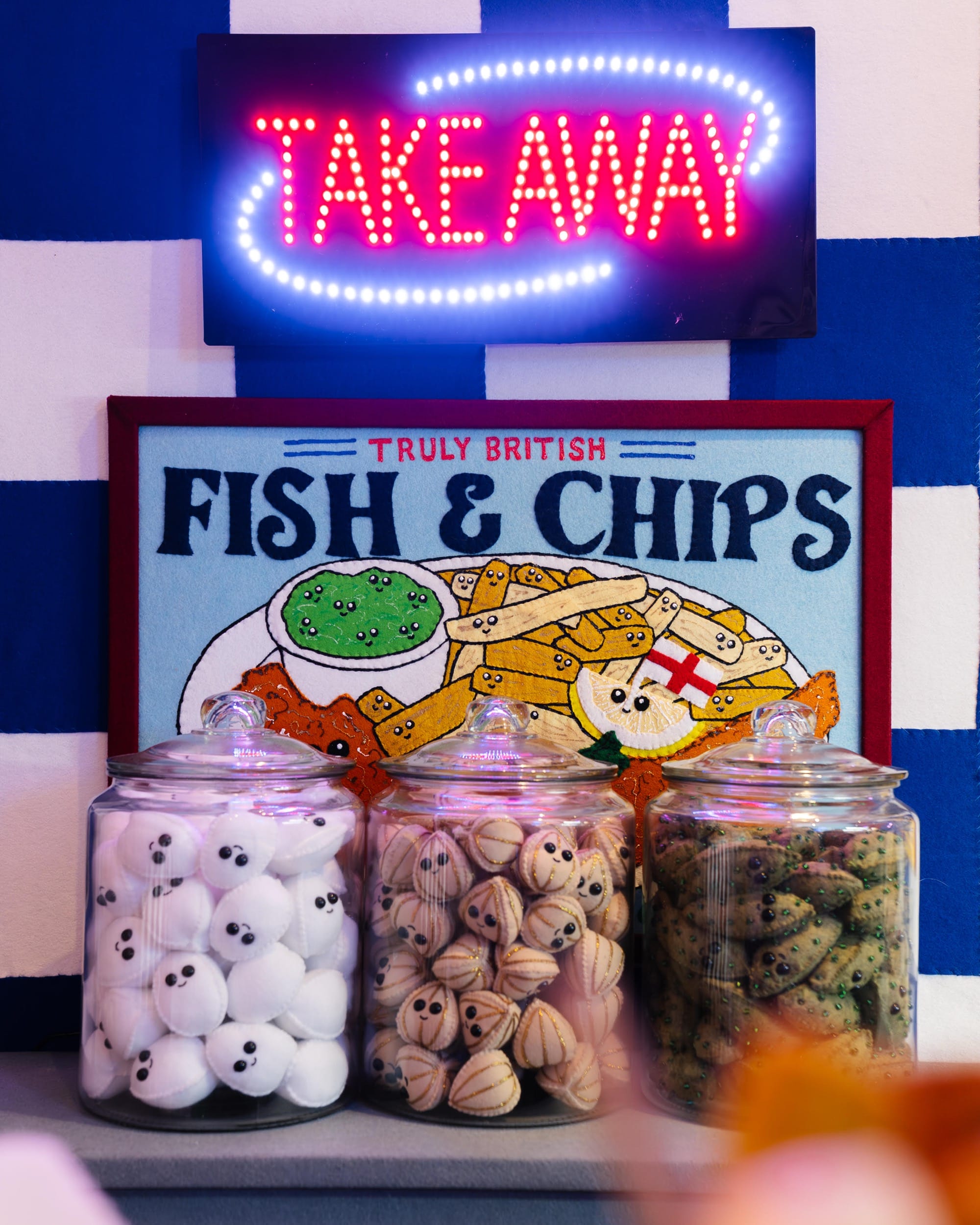 jars of pickled eggs and other veggies made of felt, under a neon sign that reads "takeaway"
