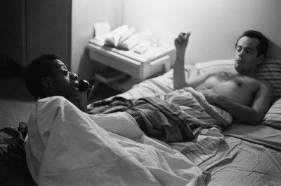 A Black mand and a white man are both lying in bed. Its avintage photo and the white man is holding a cigarette while the Black man talks on the phone, lying perpendicularly across the other man's legs.